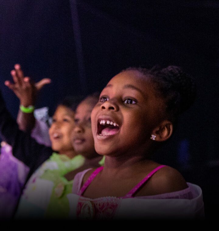 Excited children smiling, cheering, and raising their hands while watching Disney On Ice and Monster Jam performances.